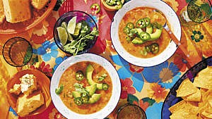 Two bowls of Mexican lentil soup topped with cilantro, avocado and jalapeños on a table with a colourful floral tablecloth alongside bowls of cornbread, cut limes and sparkling drinks
