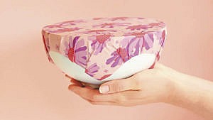 Floral patterned beeswax wrap covering half a cantaloupe in the palm of a hand against a pink background