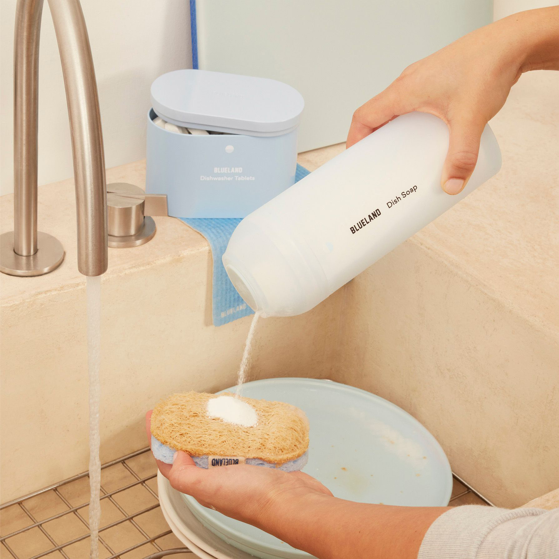 A refillable white bottle of blue land dish powder pouring onto a sponge that is washing a plate over a sink