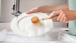A closeup of hands holding a white ceramic dish under running water coming from a faucet holding a wood scrub brush that scrubs the dish over a sink filled with soapy water
