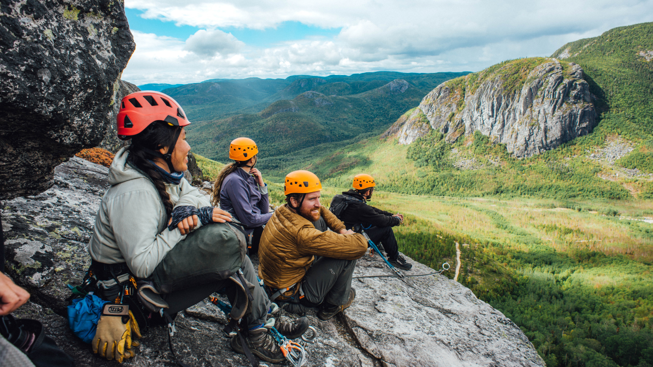 Five people in helmets sit on the side of a rock looking out into Charlevoix as part of the via ferrata climbing experience