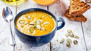 a large blue mug of butternut squash soup topped with thyme and pumpkin seeds beside toast and a spoon on a white-washed tabletop