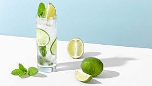 A mojito in a tall clear glass with mint leaves, lime slices and ice cubes on a white surface with a sprig of mint and a whole lime and two slices against a blue wall