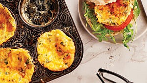 A muffin tin filled with crustless mini quiches with one removed; beside it a taupe plate with a English muffin topped with one of the quiches, a slice of tomato and argula
