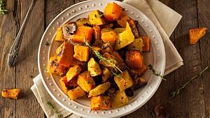A white plate holding roasted butternut squash in pieces topped with sprigs of thyme and mixed with onions on a cloth napkin on a wood table