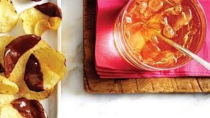 A maple rye and ginger cocktail shown from above with a spoon resting in it on a pink napkin on a cutting board on a marble table next to chocolate-dipped kettle chips