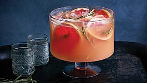 Red grapefruit punch in a glass trifle bowl with sprigs of rosemary and slices of grapefruit floating in it served in a trifle bowl with two glasses next it on a table
