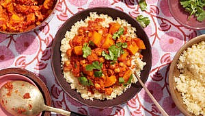 A bowl of slow cooker moroccan vegetable stew, with butternut squash, chickpeas and cilantro on a bed of couscous with a spoon inserted in the couscous to one side on a floral tablecloth with a bowl of couscous, more stews and cilantro around it
