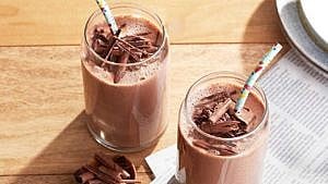two Cocoa, Banana And Tahini Breakfast Smoothies in glasses topped with chocolate shavings and with straws on a wooden table with a newspaper