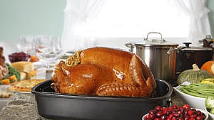 A Thanksgiving turkey that has just been removed from the oven sits in its roaster on a kitchen counter in front of a setting for a Thanksgiving dinner.