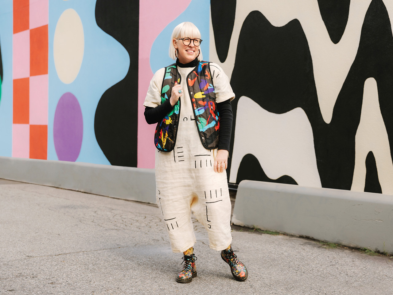 Vancouver entrepreneur Anna Heyd wearing a maximalist style outfit including a patterned white jumpsuit and a black vest with graphic shapes, photographed against a murale.