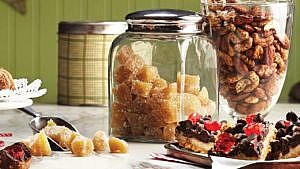 candied ginger in a glass jar with a scoop of candied ginger with teh candy spilling out onto marble counter beside other holiday treats