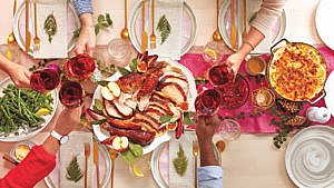 A Thanksgiving table laden with turkey, red wine, and six place settings with white plates and gold cutlery and a leaf napkin on each plate and hands reaching in to clink glasses together