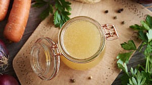 Bone broth in a glass jar on a wooden table, with root and leafy vegetables