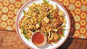 Stir-fried noodles with veggies and crispy tofu served on a plate with a side of chili oil