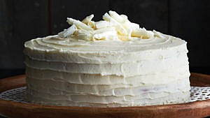 A yellow vanilla layer cake frosted with vanilla buttercream icing and topped with white chocolate shavings on a lace doily on a wooden tray