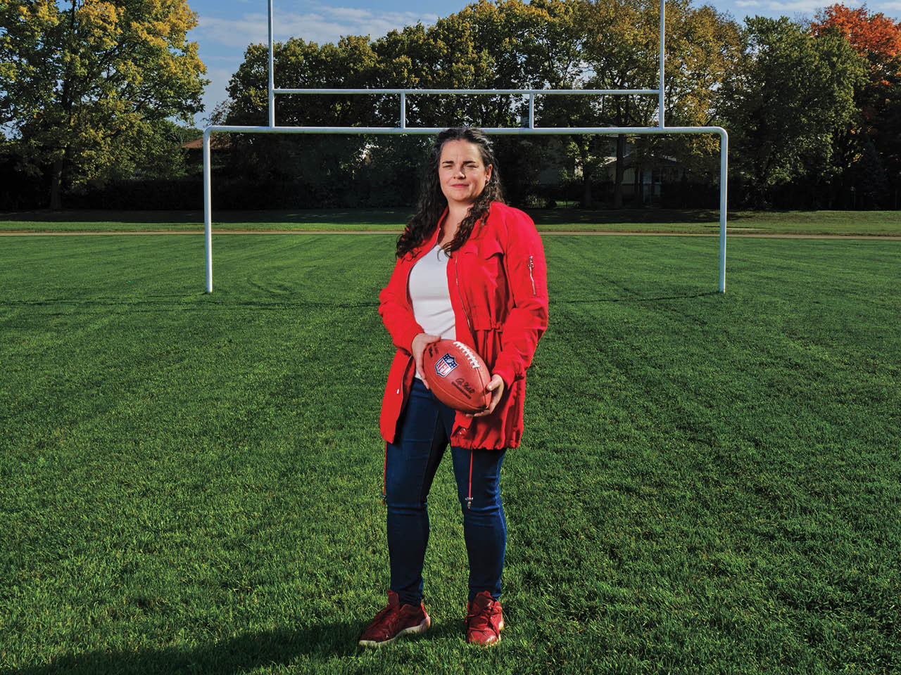 Carly Fennell, in jeans and a bright red coat, stands in a football field holding an NFL football.