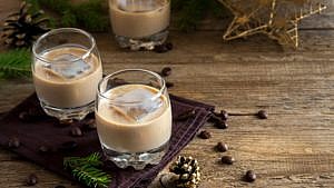 Three glasses of homemade Bailey's Irish cream liqueur on a wood table, each with a large ice cube and paired with a gold star ornament, best homemade baileys Irish cream recipe