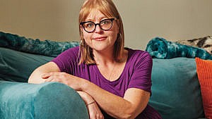 Emily Wright sits on an turquoise couch, wearing a purple T-shirt and glasses. Her arms are folded over one another on the arm of the couch. Wright has been on Ozempic since 2018.